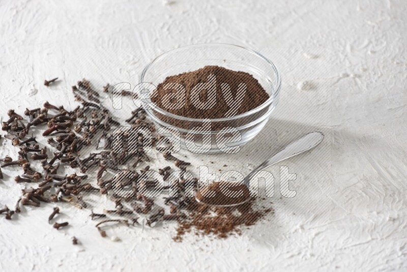 A glass bowl and a metal spoon full of cloves powder with cloves grains spread on a textured white flooring