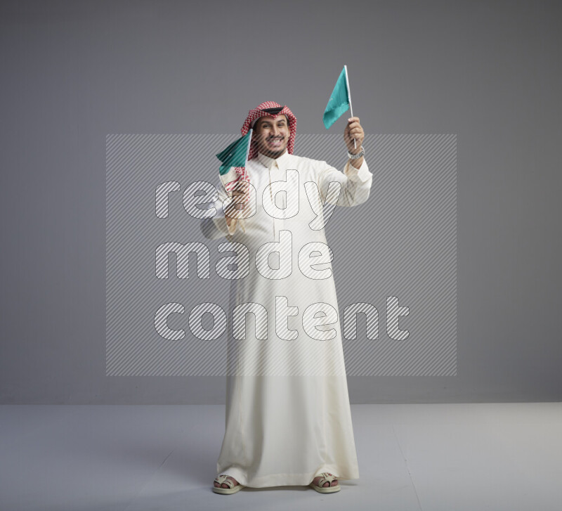 A Saudi man standing wearing thob and red shomag raising small Saudi flag on gray background