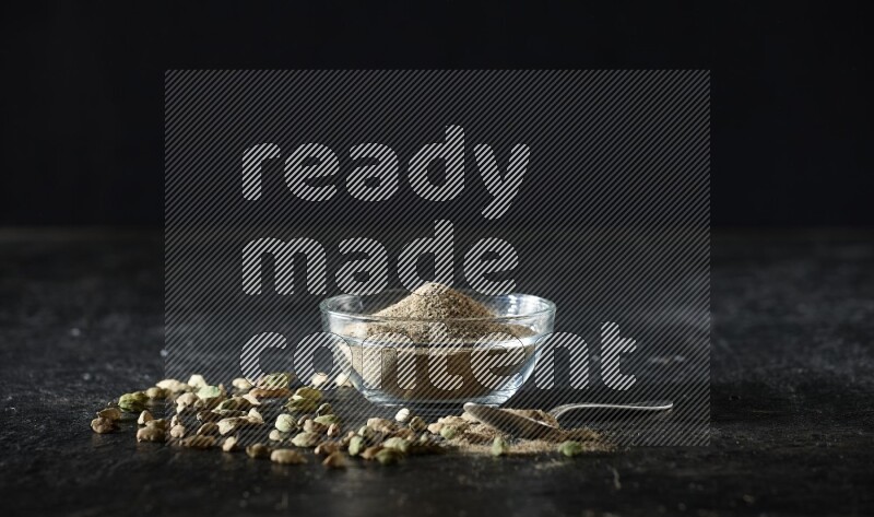 A glass bowl and a metal spoon full of cardamom powder with cardamom seeds beside them on textured black flooring
