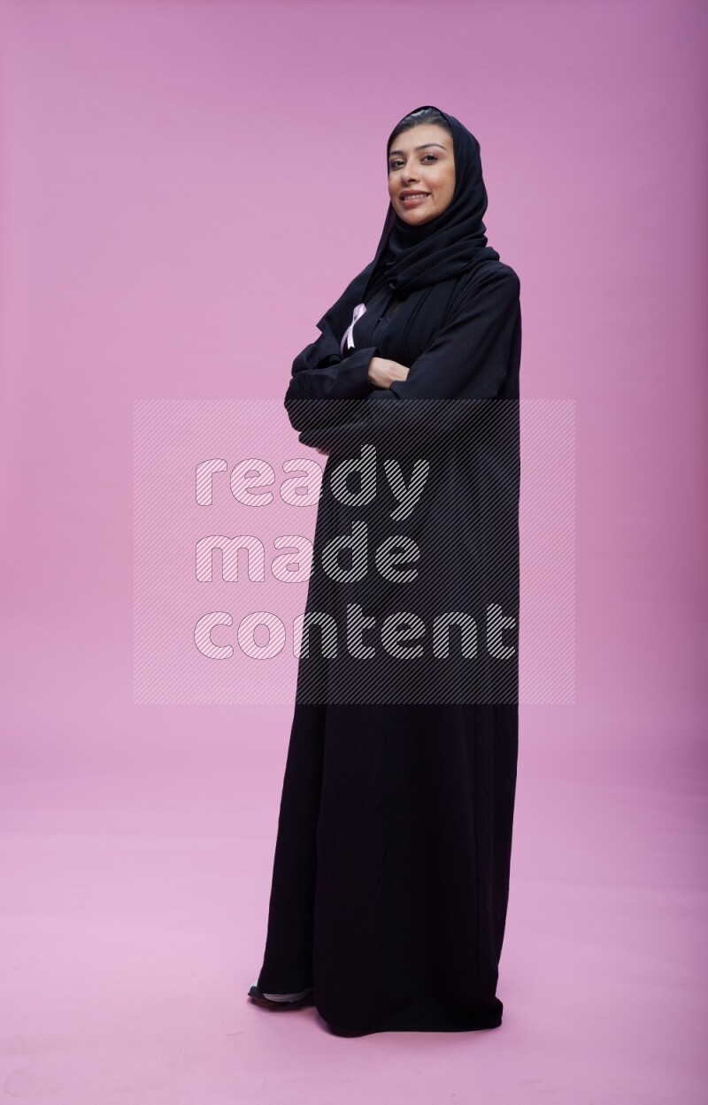 Saudi woman wearing pink ribbon on Abaya standing crossed arms on pink background