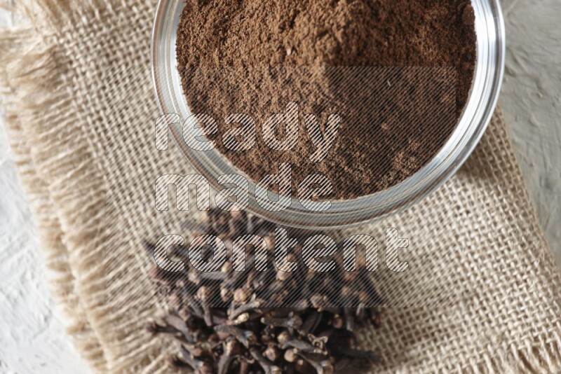 A glass bowl full of cloves powder and cloves grains on a burlap piece on a white flooring