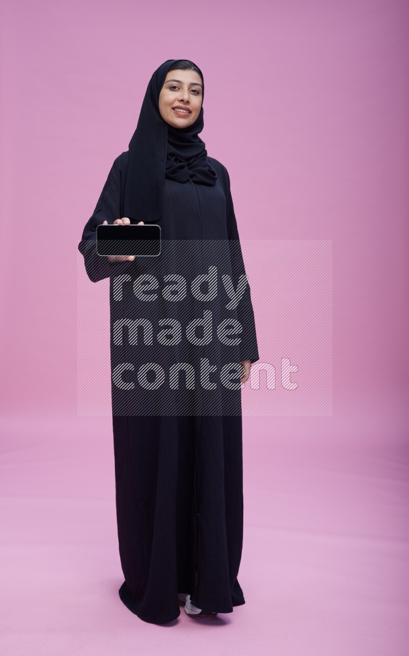 Saudi woman wearing Abaya standing showing phone to camera on pink background