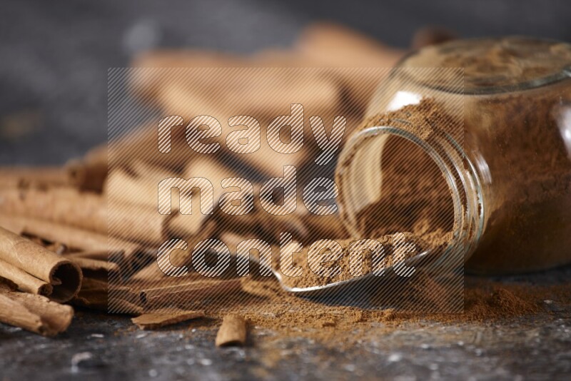 Herbal glass jar full cinnamon powder flipped and a metal spoon full of powder surrounded by cinnamon sticks on textured black background in different angles