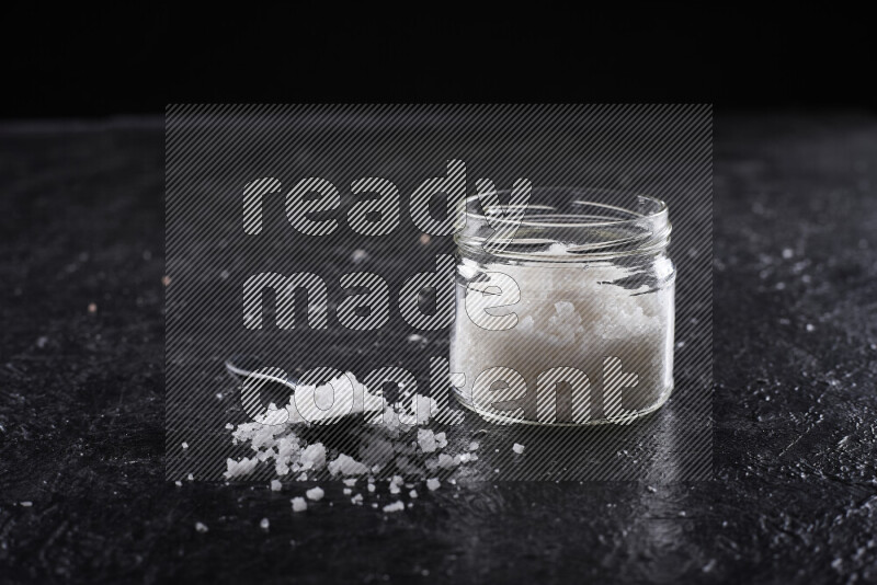 A glass jar full of coarse sea salt crystals on black background
