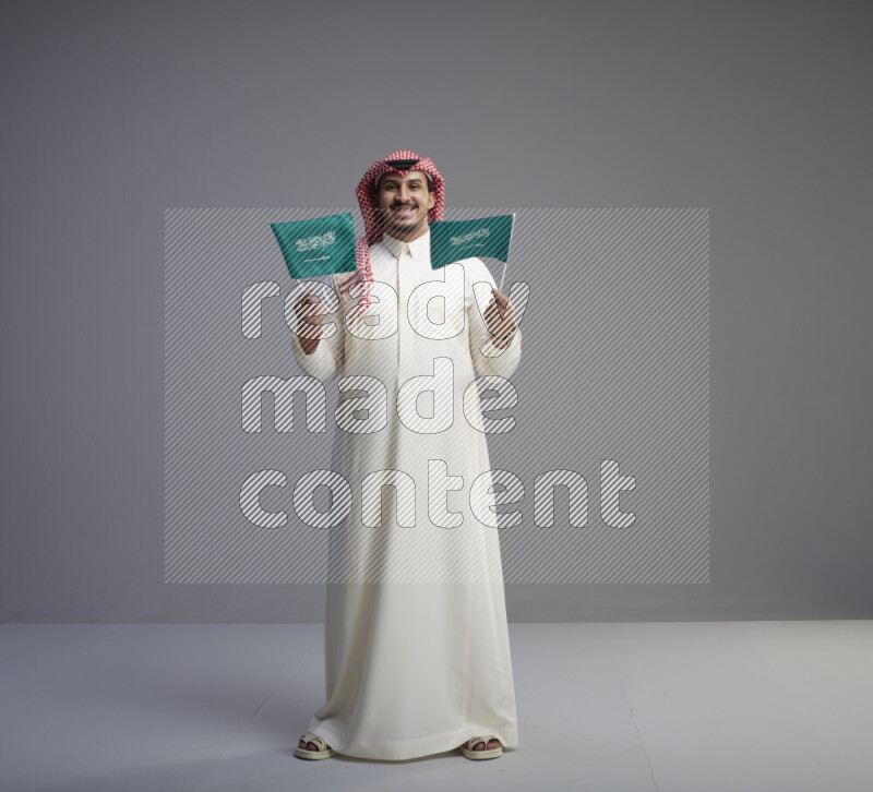 A Saudi man standing wearing thob and red shomag raising small Saudi flag on gray background