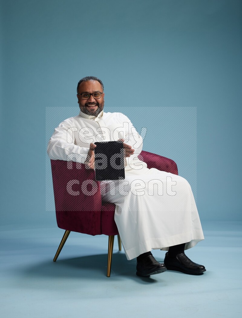 Saudi Man without shimag sitting on chair working on tablet on blue background