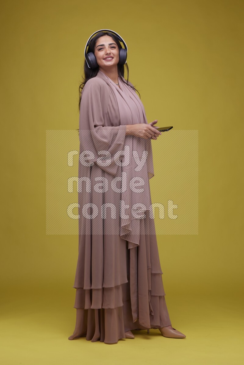 A Saudi woman Listing to Music on a Yellow Background wearing Brown Abaya