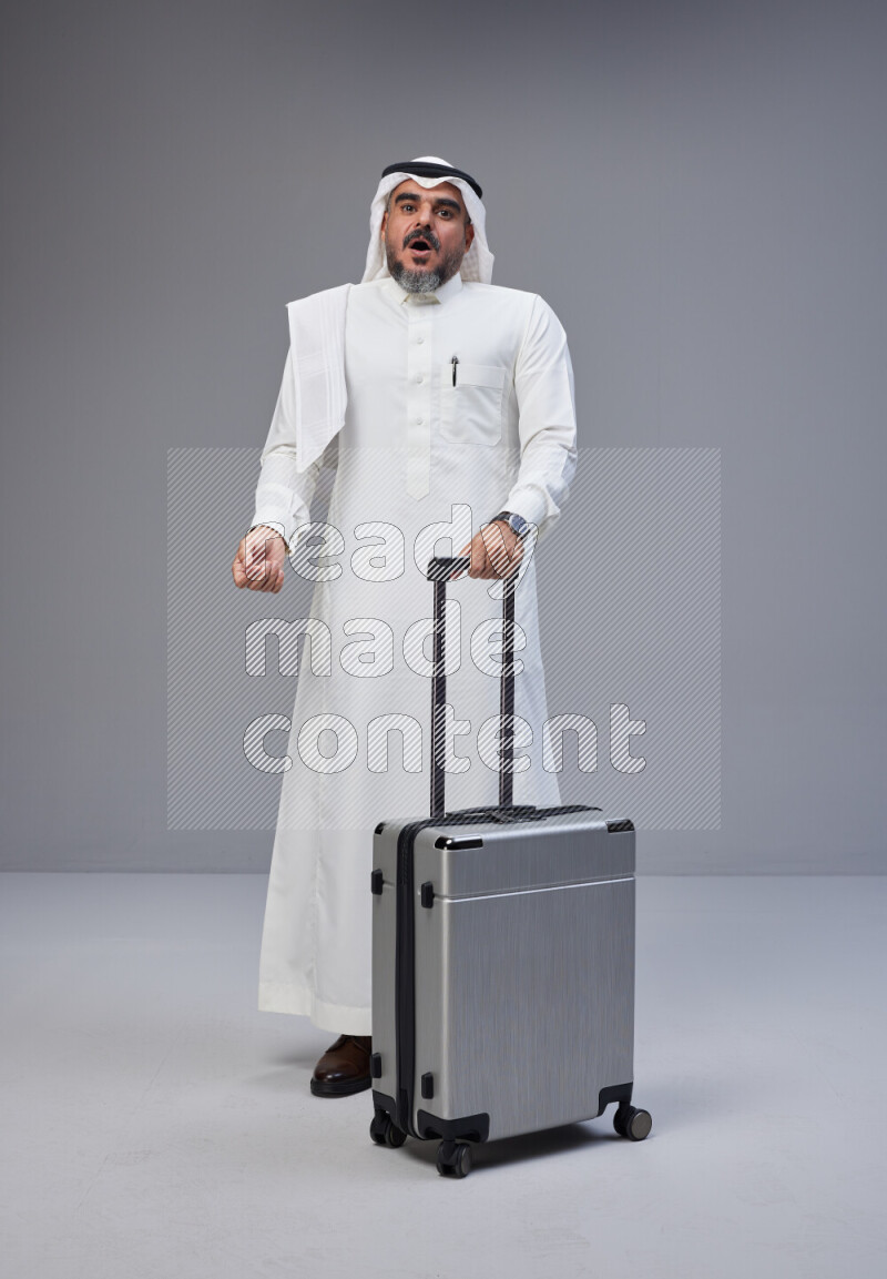 Saudi man wearing Thob and white Shomag standing holding Travel bag on Gray background