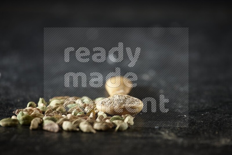 A wooden spoon full of cardamom powder and cardamom seeds next of it on textured black flooring