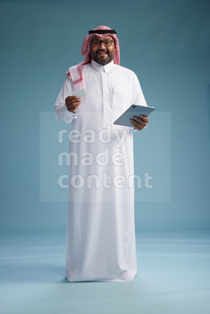 Saudi Man with shimag Standing holding ATM card while working on tablet on blue background