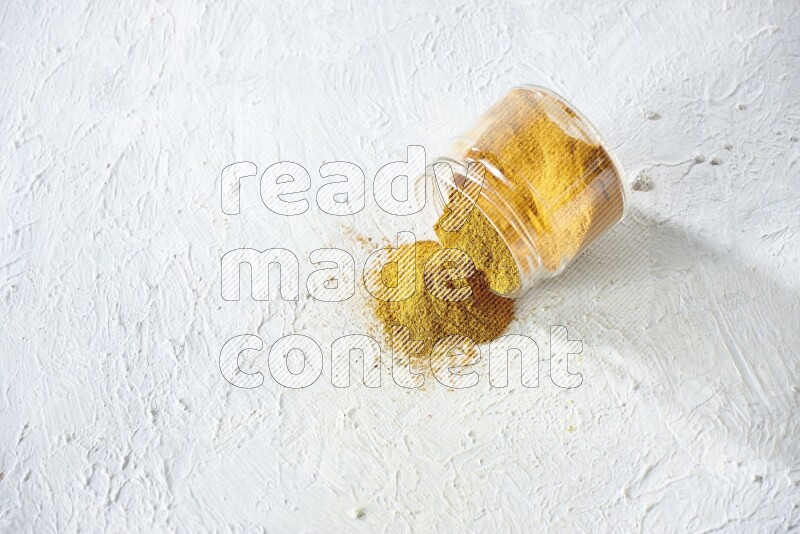 A flipped glass spice jar full of turmeric powder and powder fell out it on textured white flooring