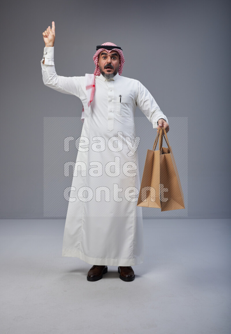 Saudi man Wearing Thob and red Shomag standing holding shopping bag on Gray background