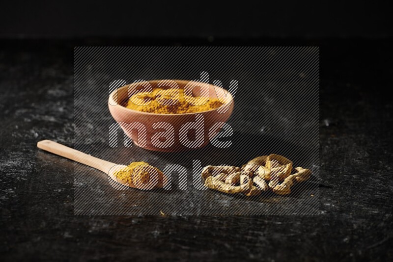 A wooden bowl and a wooden spoon full of turmeric powder with dried turmeric fingers on textured black flooring