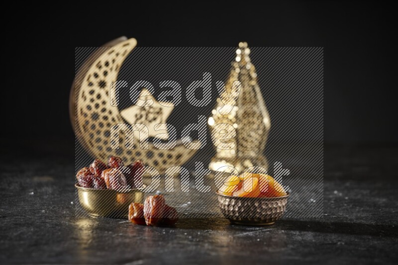 Dates in a metal bowl with dried apricot beside golden lanterns in a dark setup