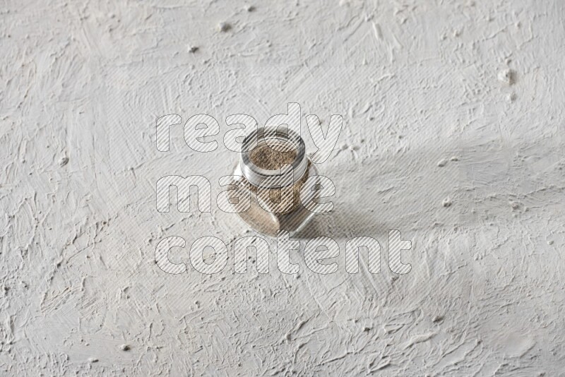 A glass spice jar full of powder black pepper on textured white flooring