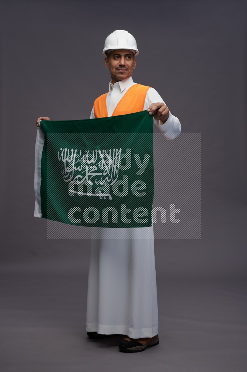 Saudi man wearing thob with engineer vest standing holding Saudi flag on gray background