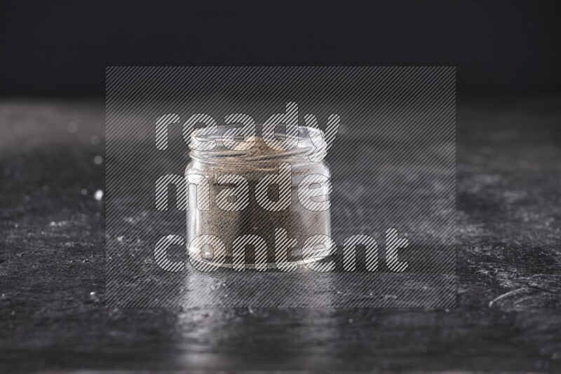 A glass jar full of black pepper powder on a textured black flooring