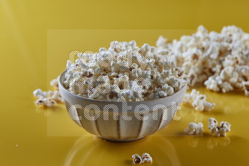 A white pottery bowl full of popcorn with popcorn beside it on a yellow background in different angles