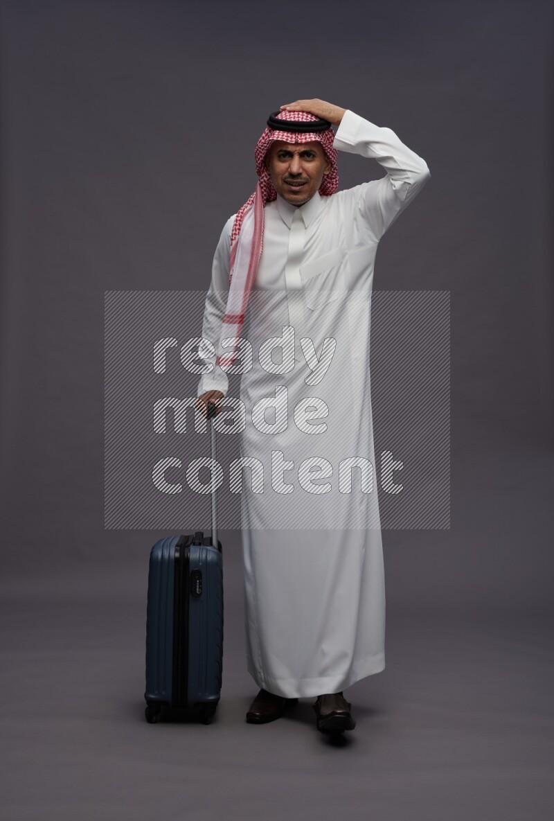 Saudi man wearing thob and shomag standing holding bag on gray background