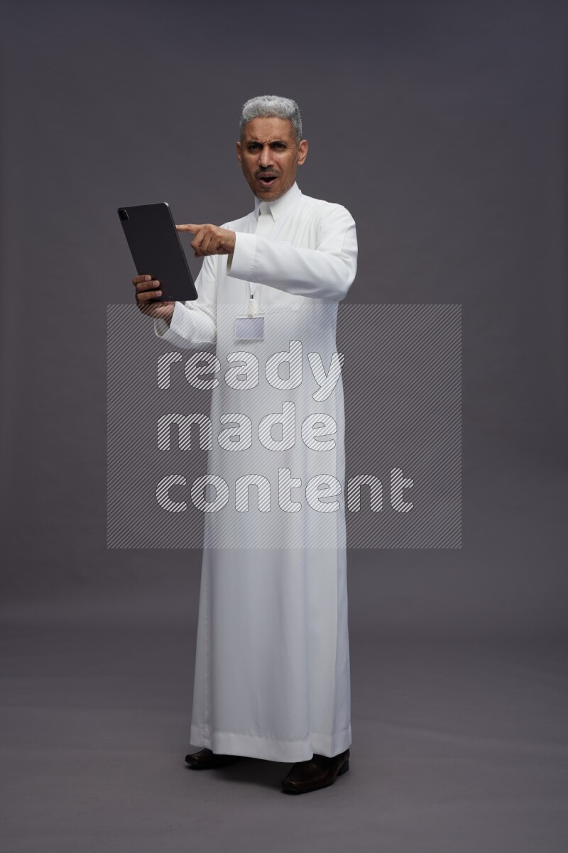 Saudi man wearing thob with neck strap employee badge standing working on tablet on gray background