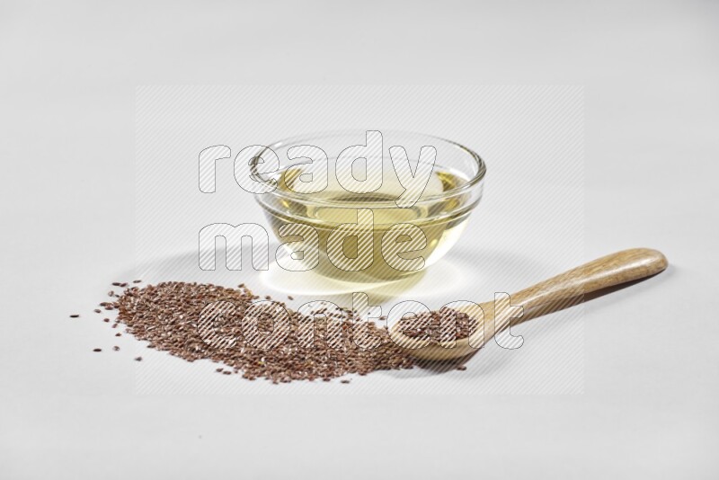 A glass bowl full of flaxseeds oil and wooden spoon full of flaxseeds with spread seeds on a white flooring