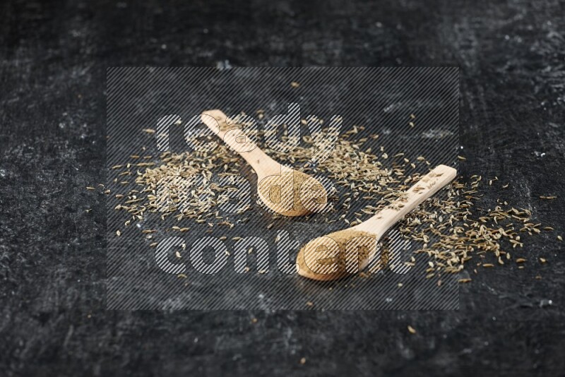 2 wooden spoons full of cumin powder with spreaded seeds on textured black flooring
