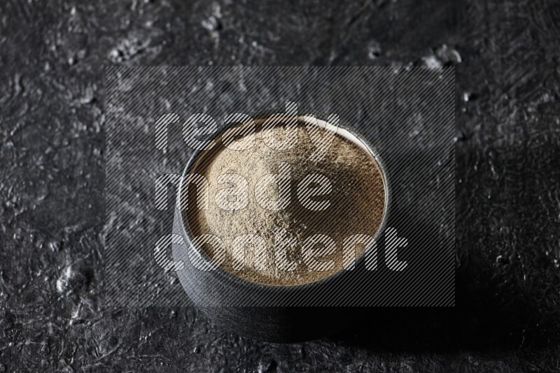 Black pottery bowl full of white pepper powder on textured black flooring