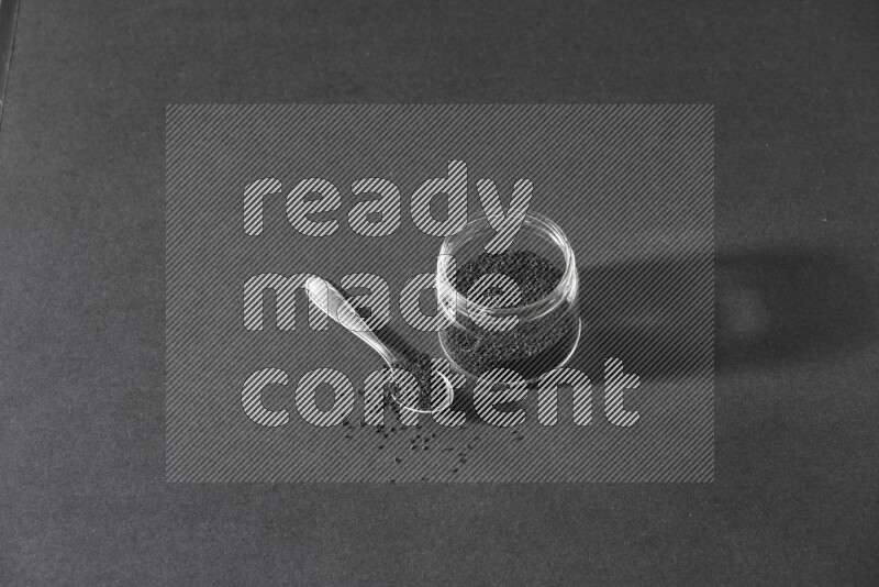 A glass jar full of black seeds with a metal spoon full of the seeds on a black flooring