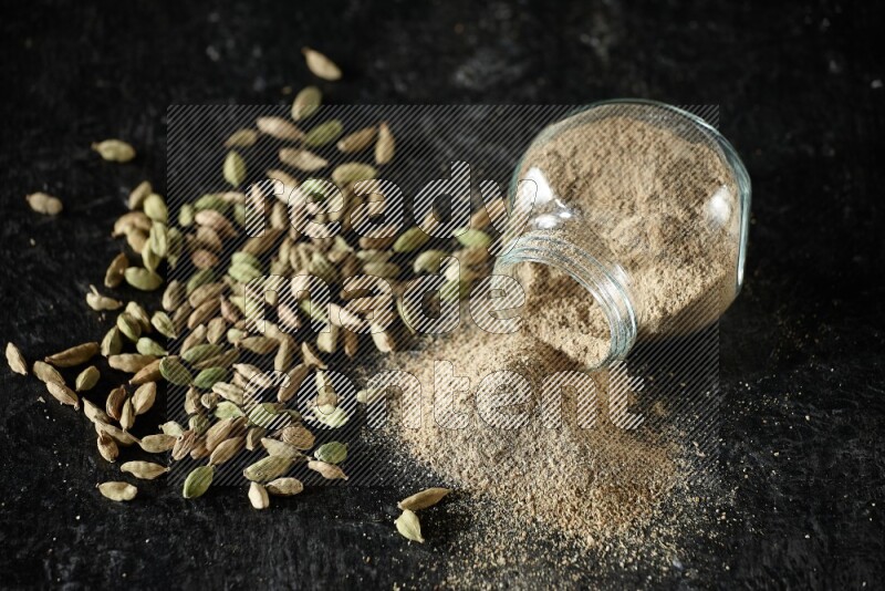 A flipped glass spice jar full of cardamom powder and powder spilled out of it with cardamom seeds spreaded on textured black flooring