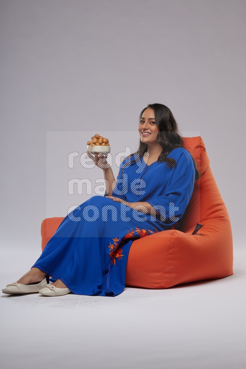 A Woman sitting on an orange beanbag wearing Jalabeya holding a plate of luqaimat