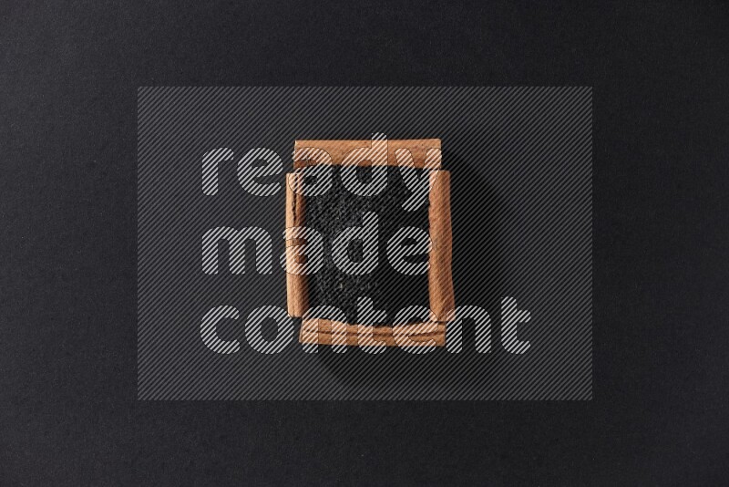 أربعة من أعواد القرفة مكونة شكل مربع بداخلها حبة البركة علي خلفية سوداء