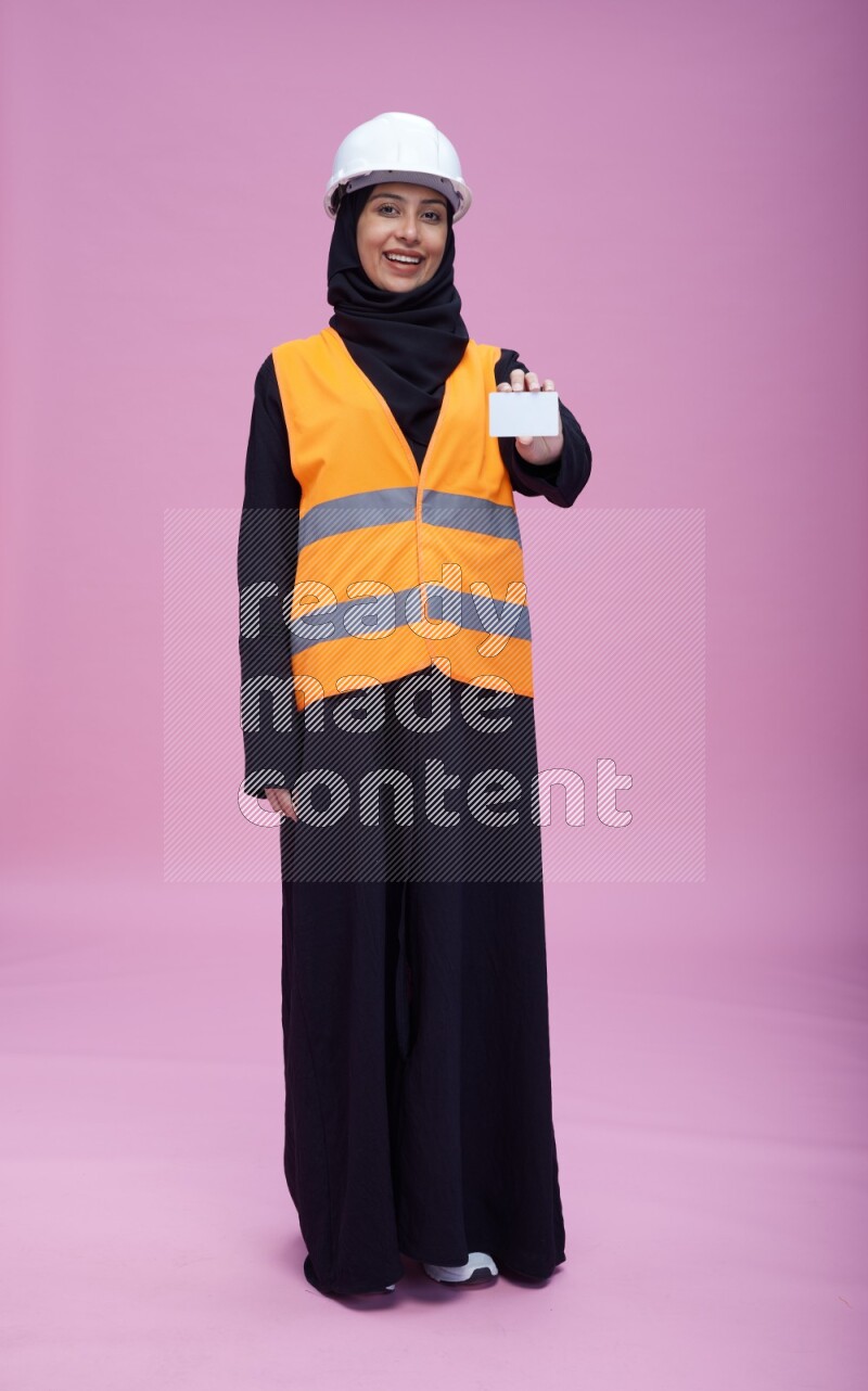Saudi woman wearing Abaya with engineer vest and helmet standing holding ATM card on pink background