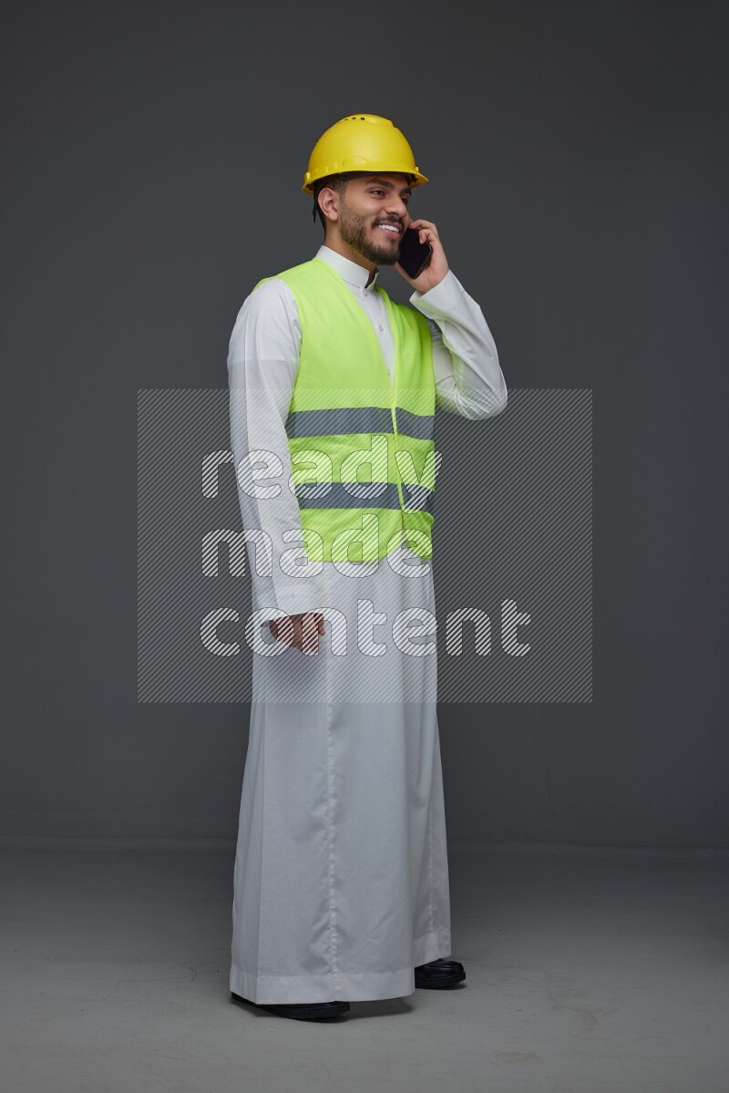 A Saudi man wearing Thobe with a yellow safety vest and white helmet standing and talking in the phone eye level on a gray background