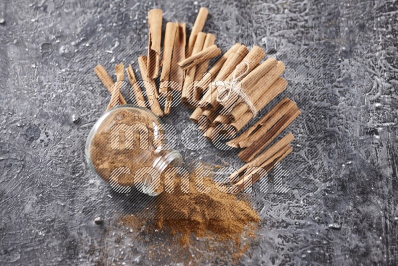 Herbal glass jar full of cinnamon powder flipped with cinnamon sticks stacked and bounded on a textured black background