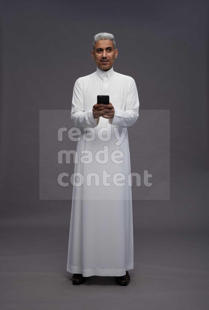Saudi man wearing thob standing texting on phone on gray background
