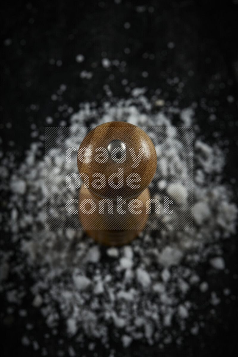 A wooden grinder standing upright and surrounded by coarse white sea salt on black background