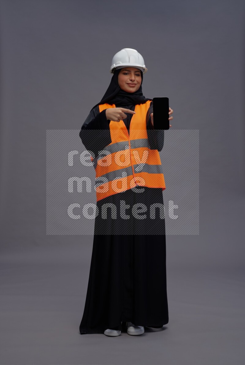 Saudi woman wearing Abaya with engineer vest standing showing phone to camera on gray background