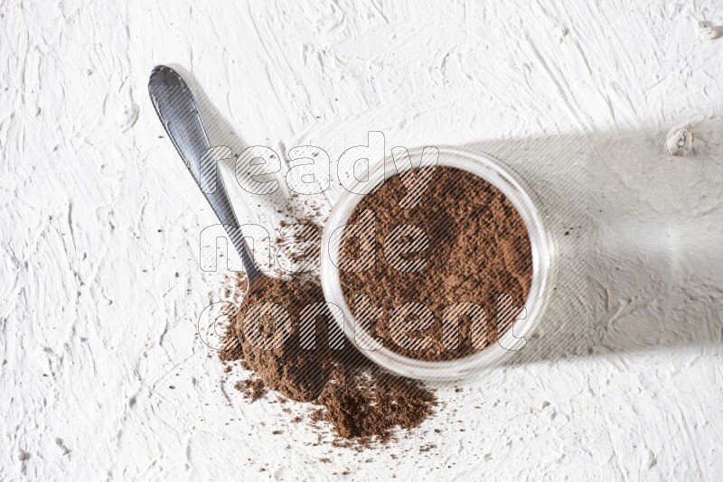 A glass jar full of cloves powder with a metal spoon on a textured white flooring