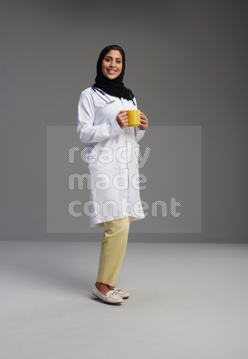Saudi woman wearing lab coat with stethoscope standing holding mug on Gray background