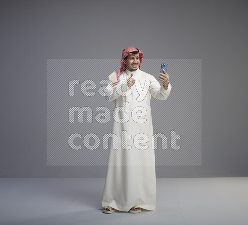 A Saudi man standing wearing thob and red shomag taking selfie on gray background