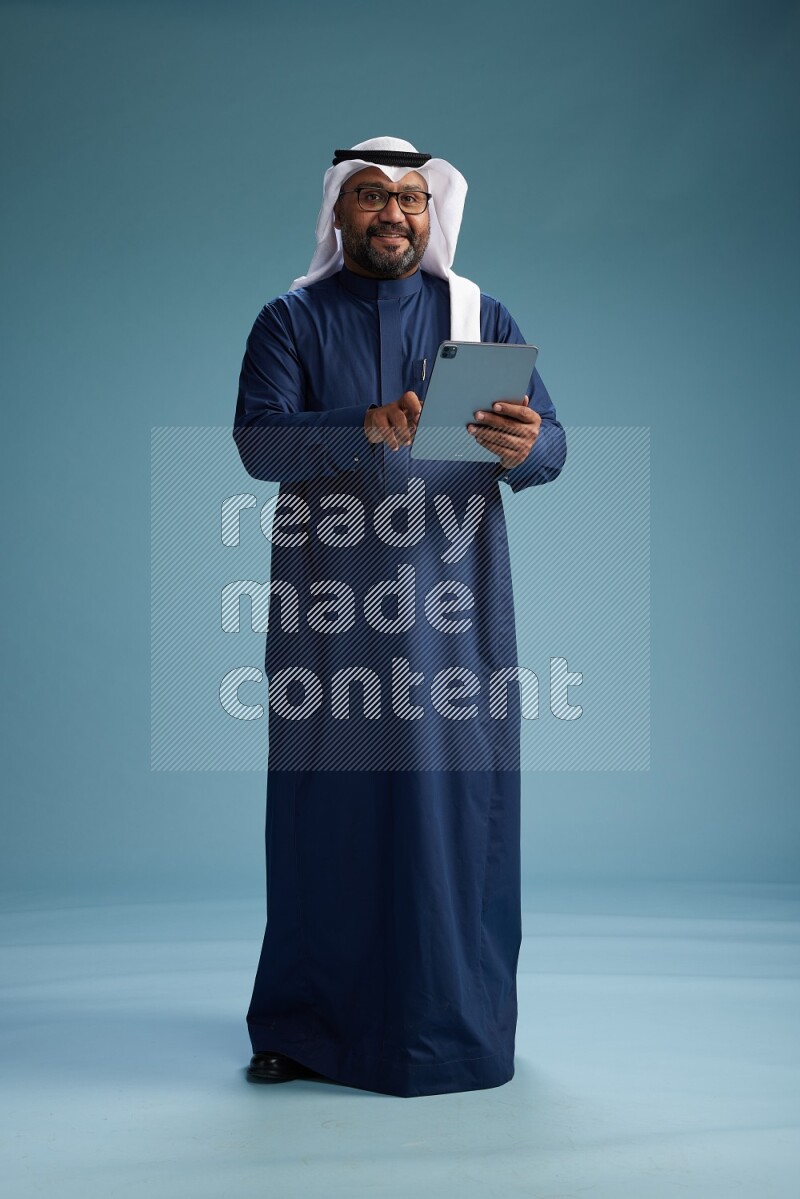 Saudi Man with shimag Standing holding ATM card while working on tablet on blue background