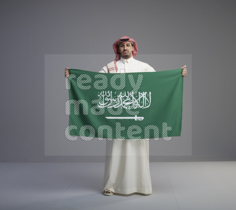 A saudi man standing wearing thob and red shomag holding big saudi flag on gray background