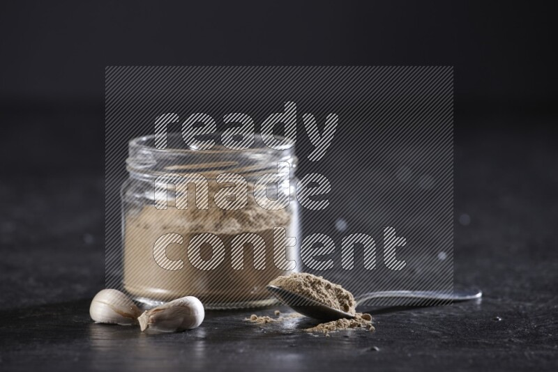 A glass jar full of garlic powder with a metal spoon full of the powder on a textured black flooring
