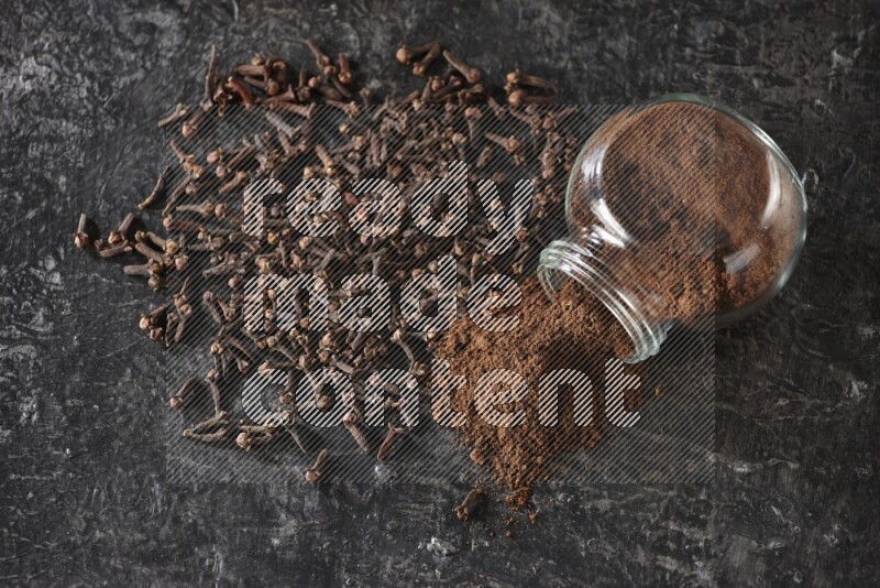 وعاء زجاجي للبهارات مقلوب ممتلئ ببودرة القرنفل والبودرة سقطت منه مع حبوب القرنفل المتناثرة على أرضية سوداء