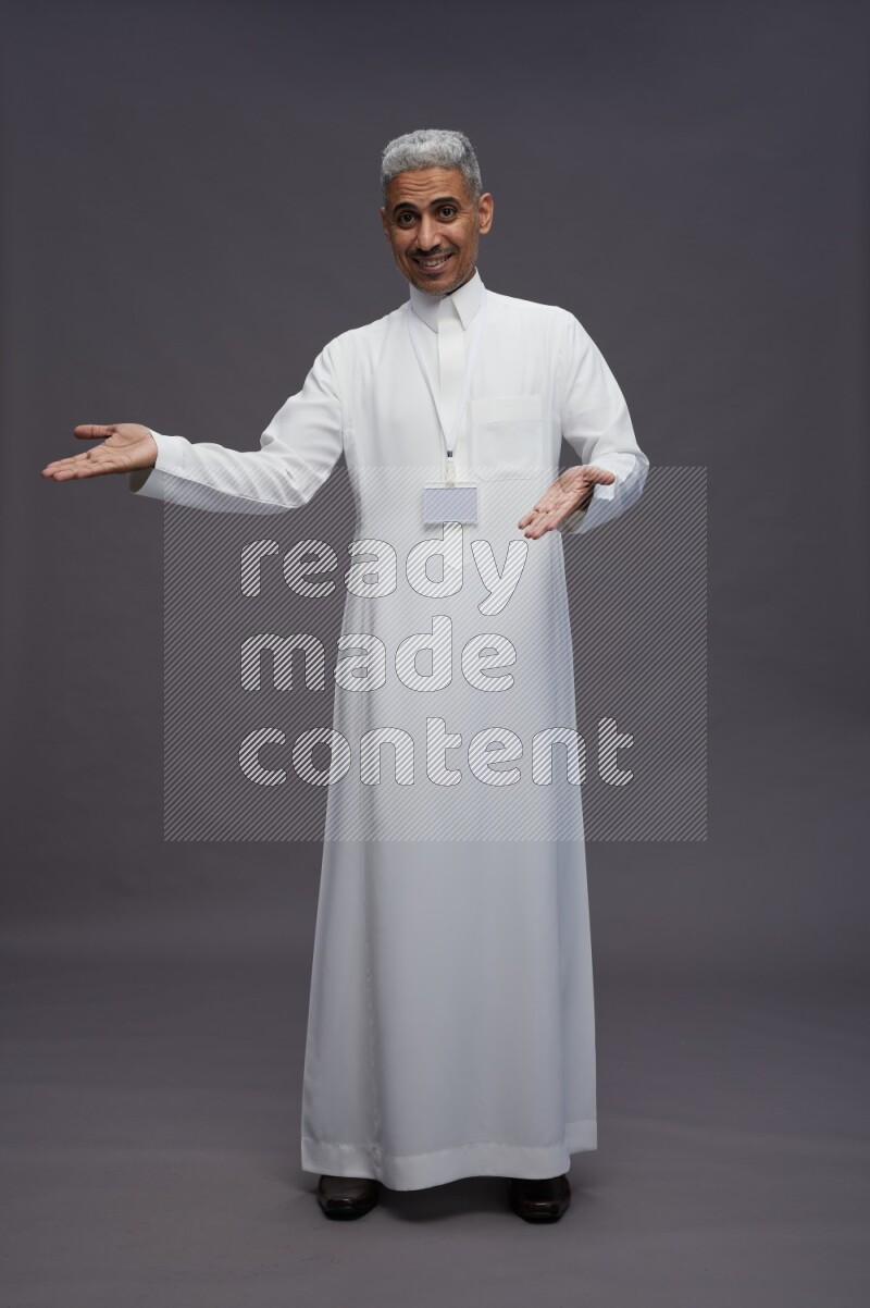 Saudi man wearing thob with neck strap employee badge standing interacting with the camera on gray background
