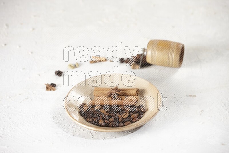 Beige plate full of coffee beans, cinnamon sticks and star anise with a coffee grinder, coffee beans, cinnamon pieces and cardamom next of it on white background