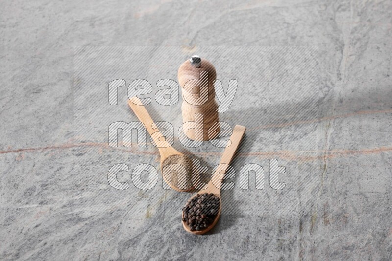 2 wooden spoons, one full of black pepper powder and the other full of black pepper beads and wooden grinder on a marble flooring