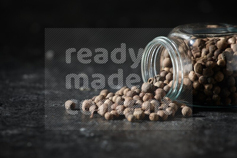 A flipped glass spice jar full of allspice whole balls and the balls spilled out of it on a textured black flooring