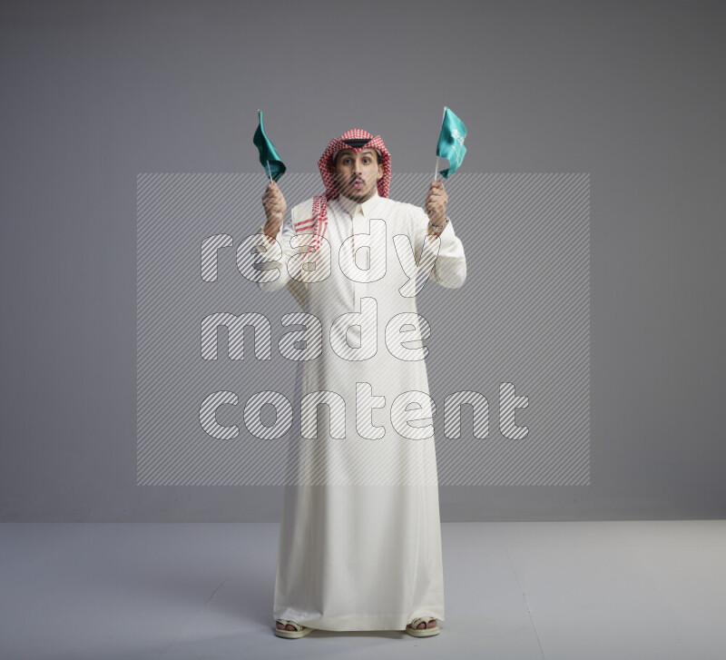A Saudi man standing wearing thob and red shomag raising small Saudi flag on gray background