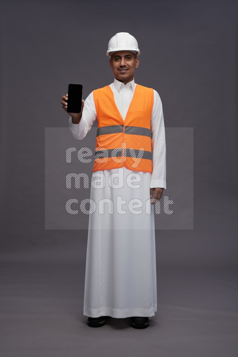 Saudi man wearing thob with engineer vest standing showing phone to camera on gray background